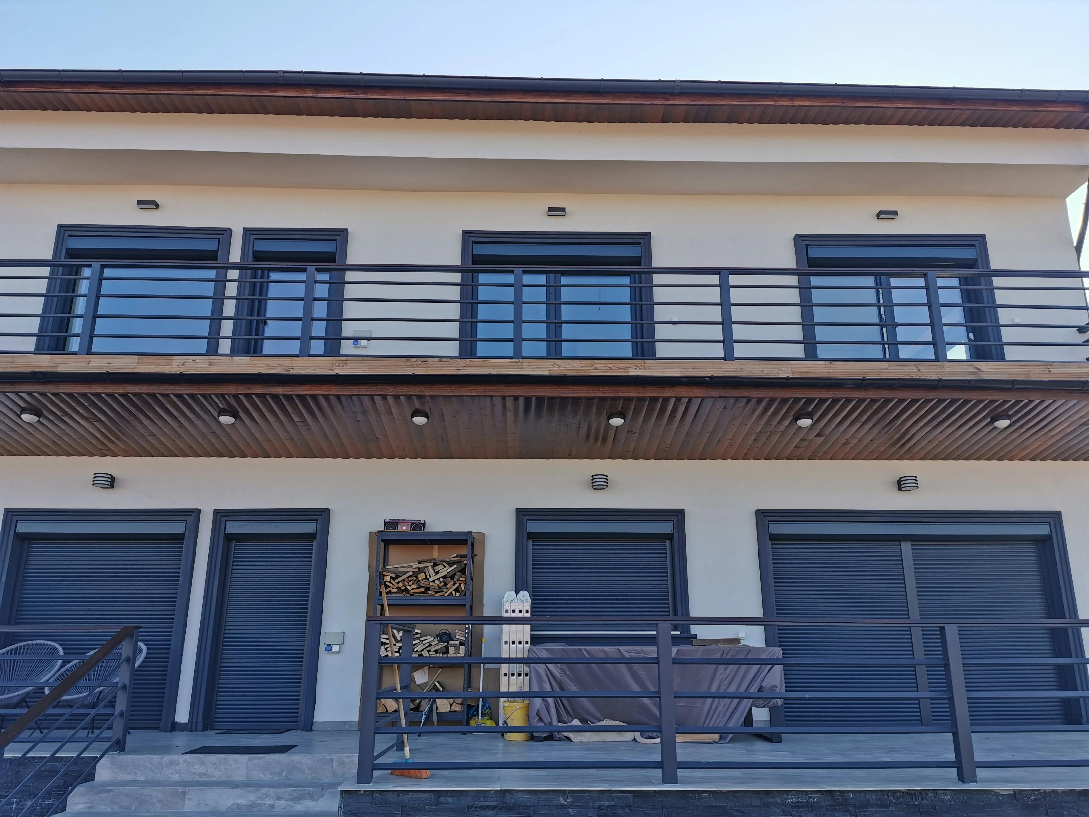 Fațadă de casă cu etaj, cu rulouri exterioare din aluminiu antracit la ferestrele de la parter, terasă cu mobilier acoperit și un raft cu lemne de foc lângă balustradă.